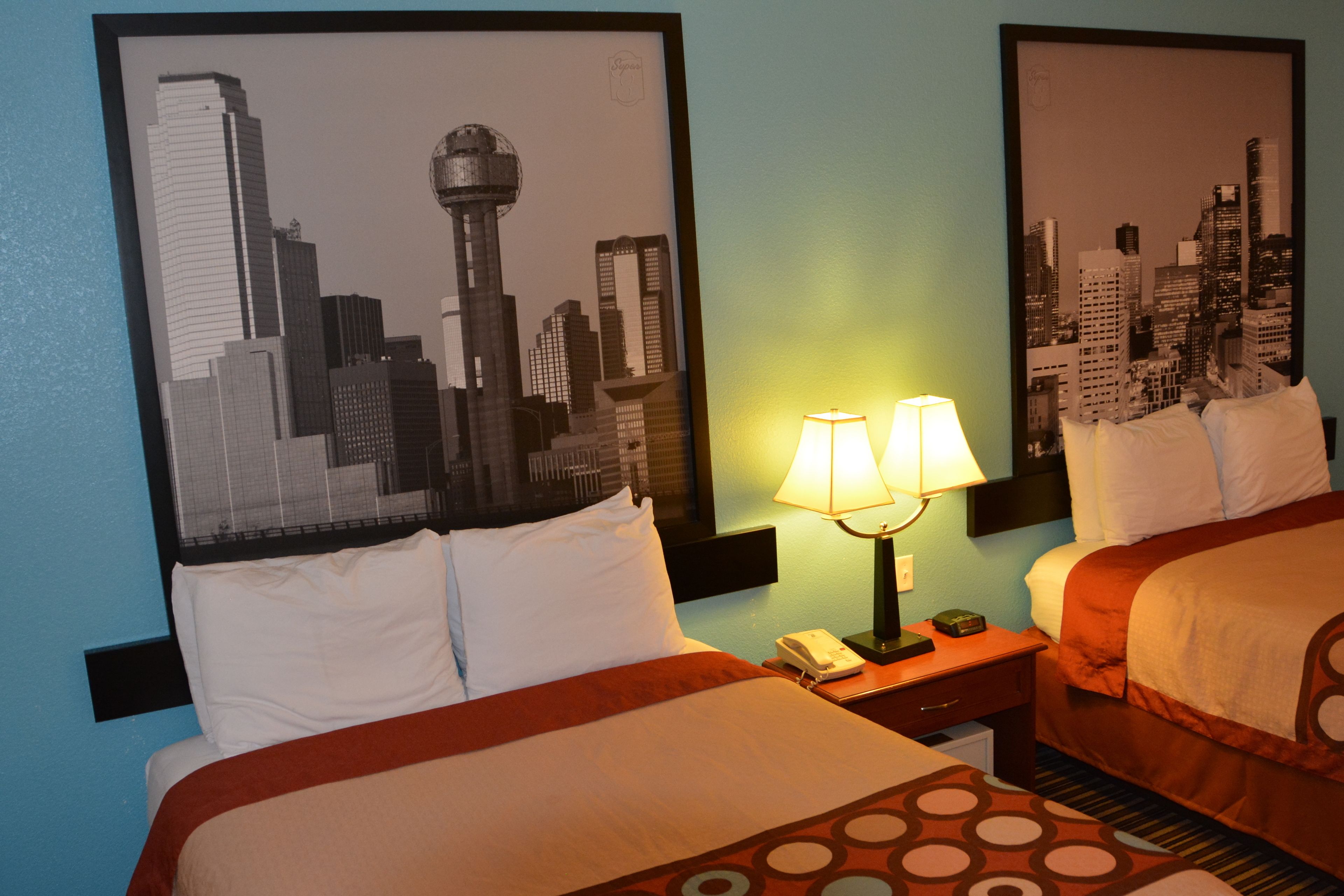 A hotel room with two beds, a cityscape artwork on the wall, a nightstand with a lamp, and a phone.