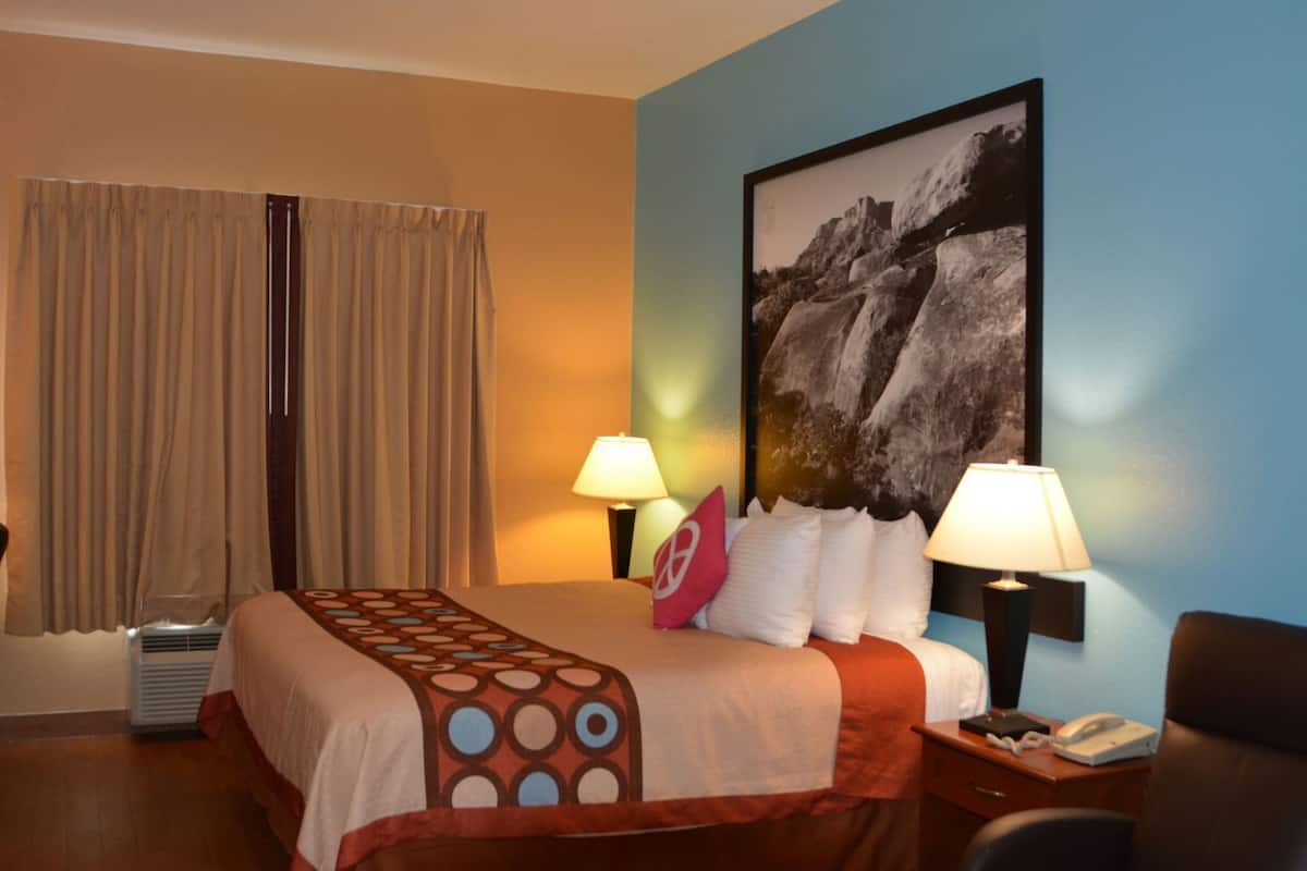 Habitación de hotel con cama, dos lámparas de noche, un cuadro grande en la pared y una ventana con cortinas.