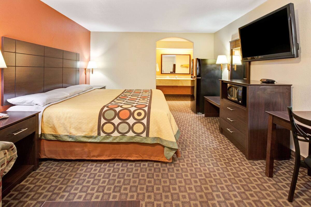 A hotel room with a large bed, a desk with a chair, a television, a microwave, and a bathroom visible through an archway.