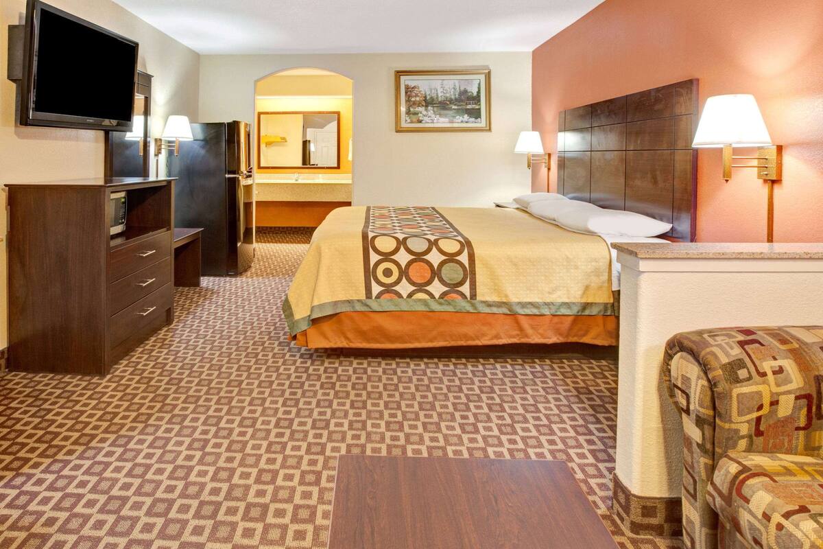 A hotel room with a bed, a TV, a desk, a chair, and a bathroom visible through an archway.