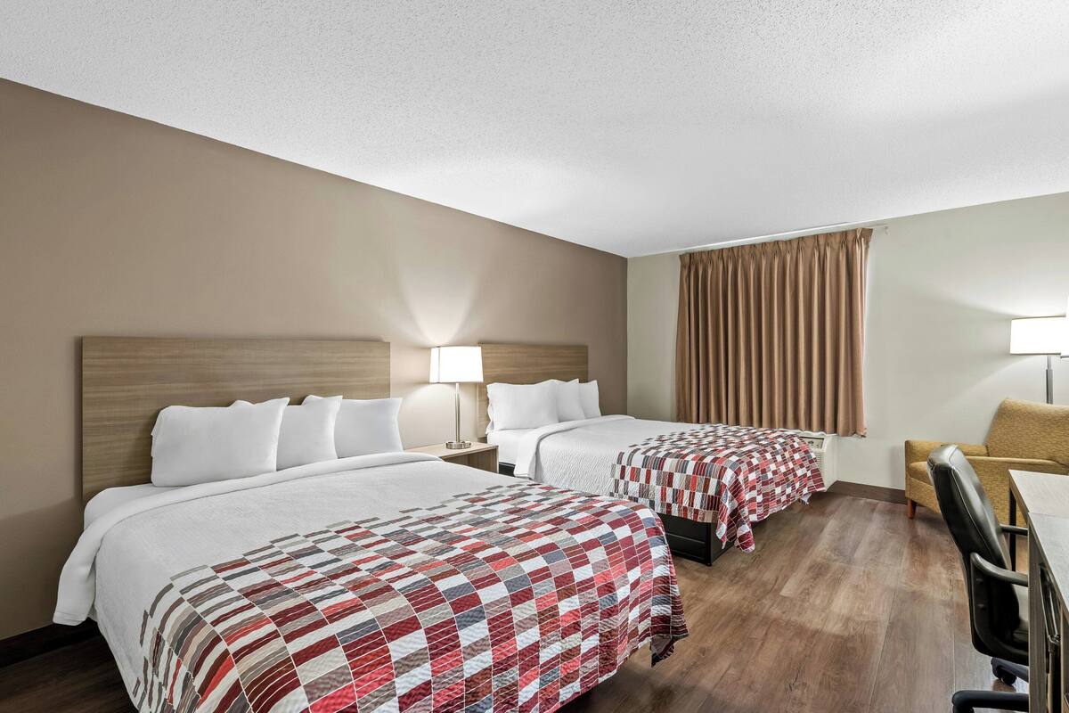 Een hotelkamer met twee bedden, een bureau, een stoel, een lamp en een raam met gordijnen.