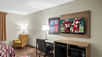 A hotel room with a bed, a desk, a chair, a lamp, a TV, and a microwave.