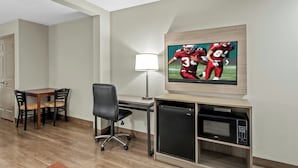 A hotel room with a wooden desk, a black office chair, a television mounted on the wall, a lamp, and a built-in microwave.