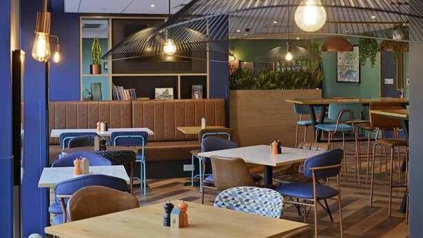 Interior de restaurante moderno com mesas de madeira e cadeiras azuis, área com bancos de madeira e luminárias pendentes.