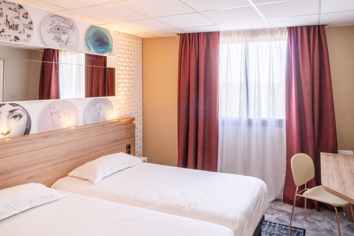 A hotel room with two beds, a wooden headboard, a desk with a chair, a large mirror, and artwork on the wall.
