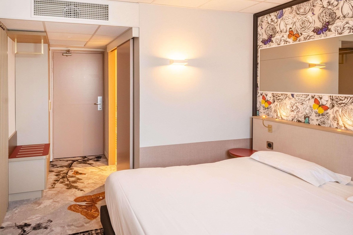 Una habitación de hotel con una cama, un espejo con papel tapiz floral, una mesita de noche y una puerta que da a otra habitación.