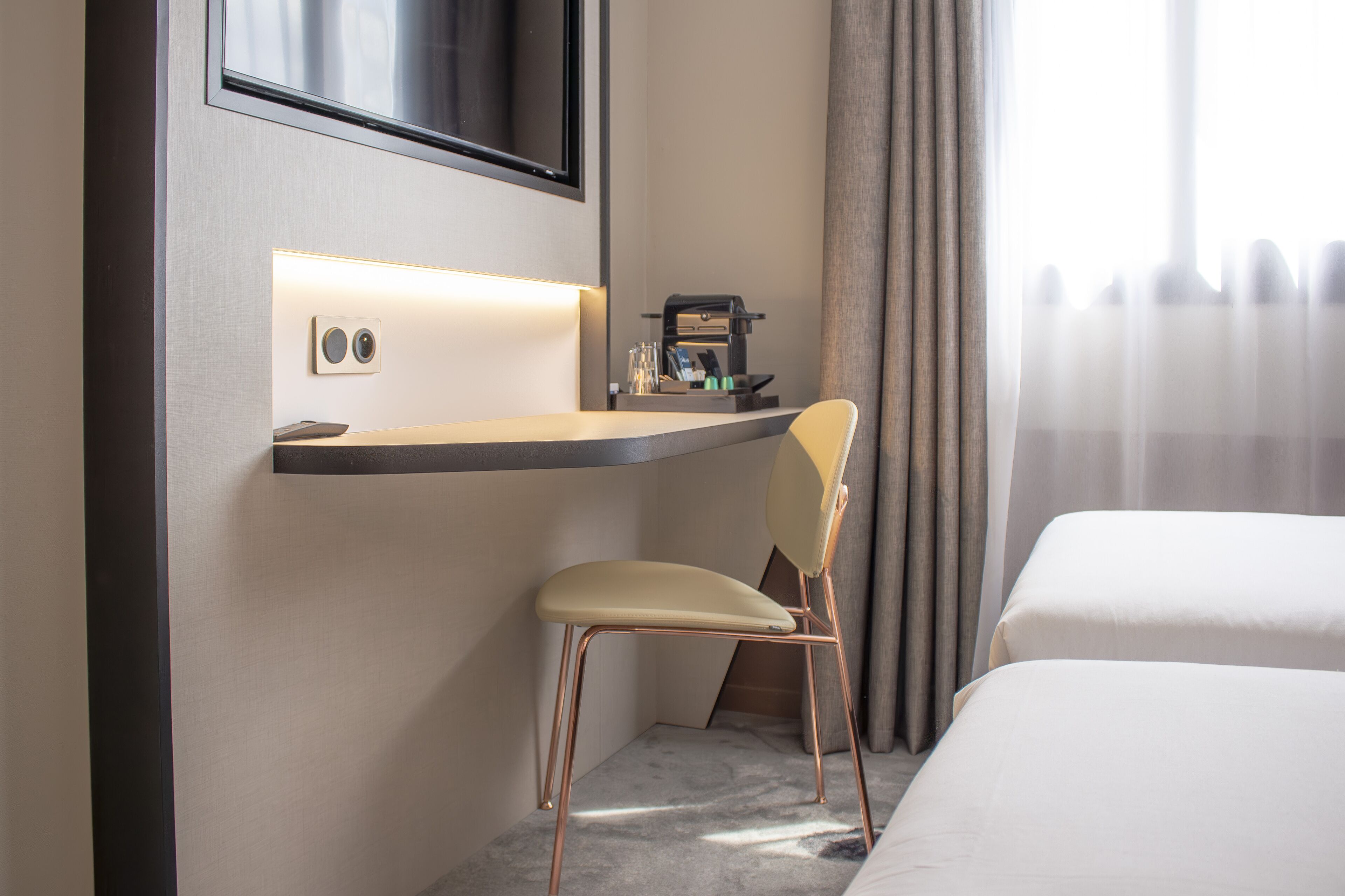 A hotel room with a bed, a desk with a chair, a coffee machine, and a TV mounted on the wall.