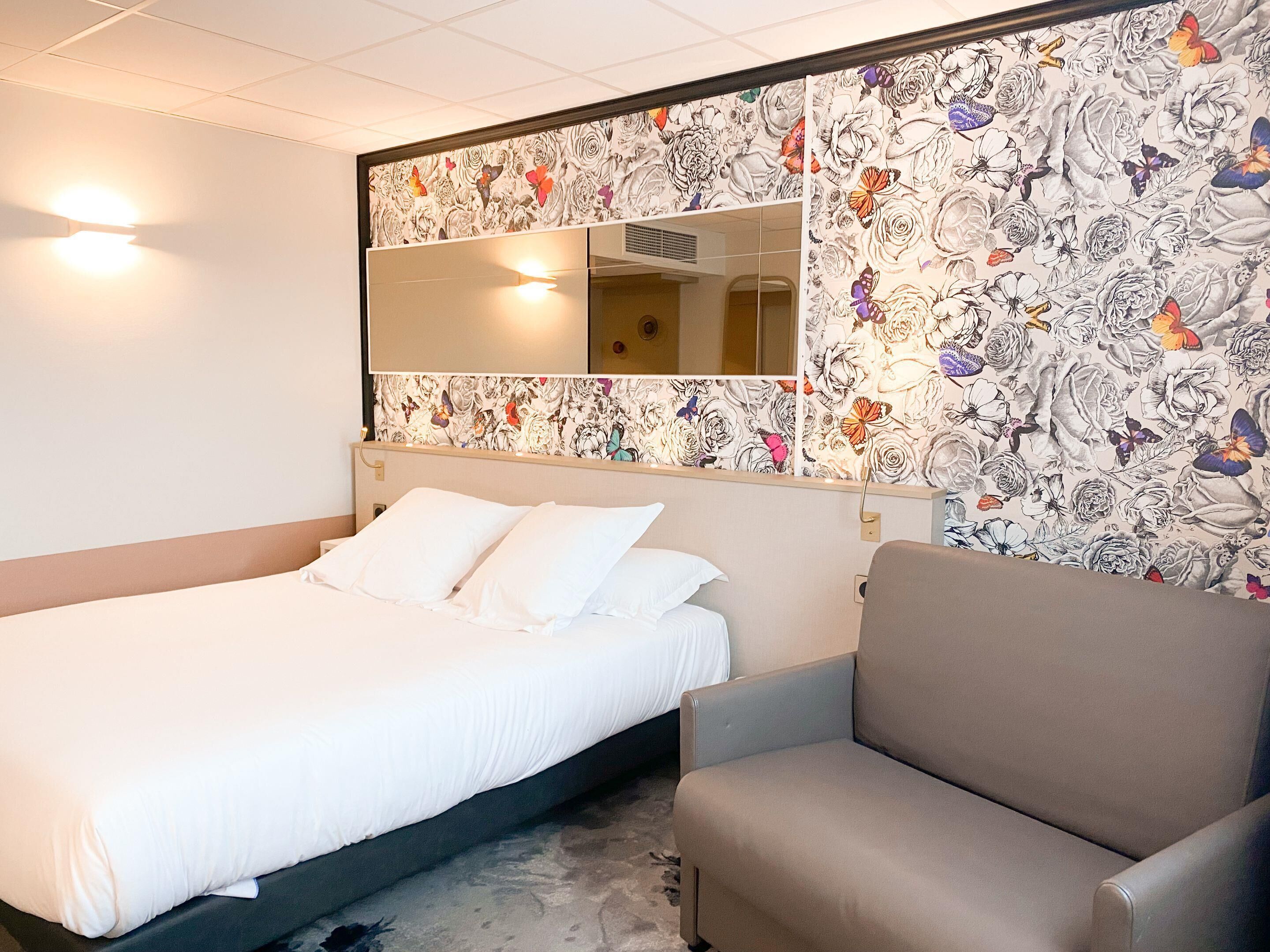 A hotel room with a bed, a chair, a large mirror, and floral wallpaper.
