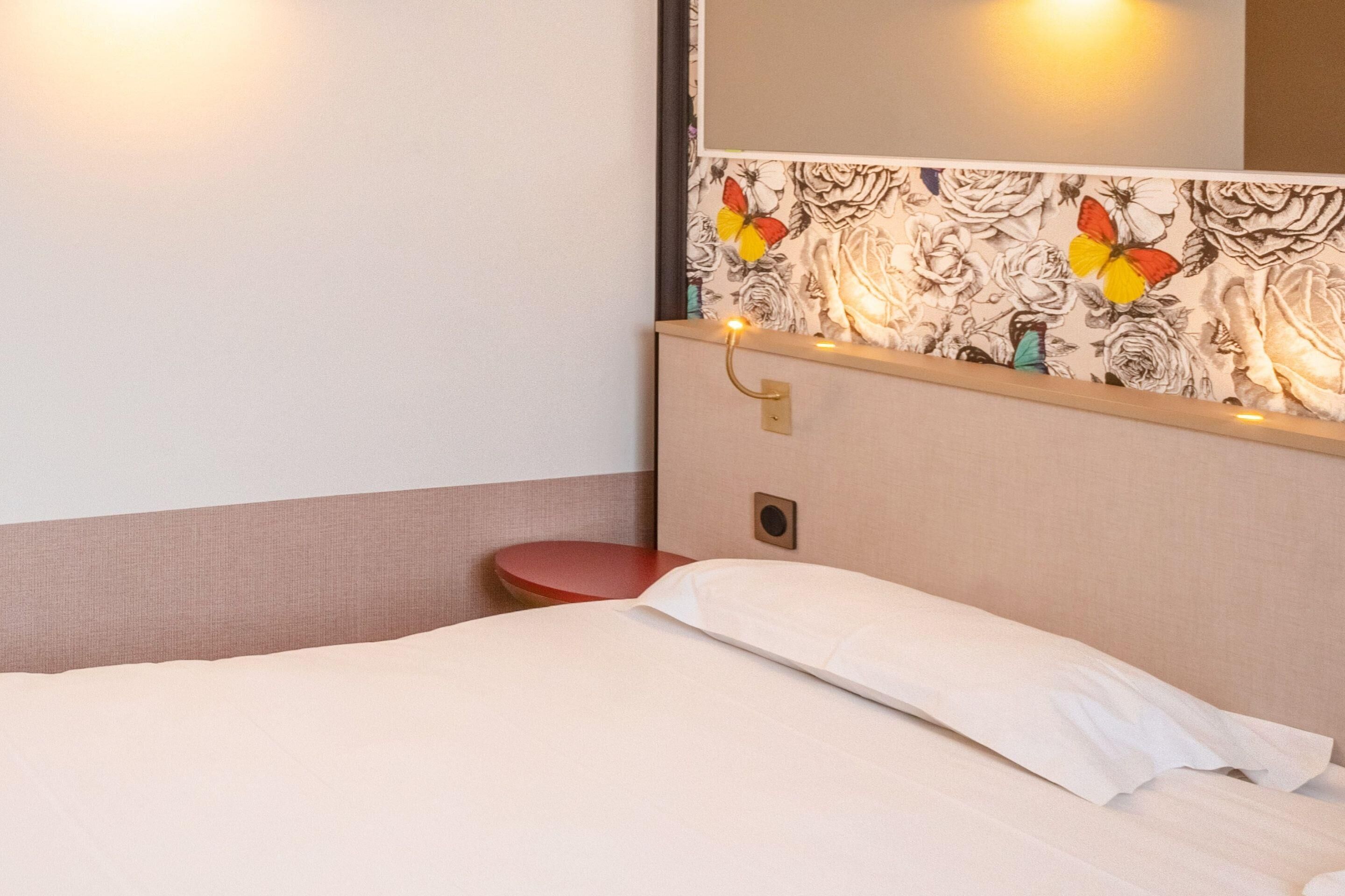 Ein Hotelzimmer mit einem Bett, einem roten Sessel, einem Wandspiegel und einem Blumenmuster an der Tapete.