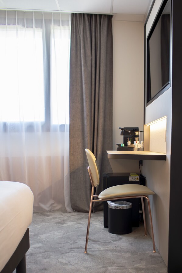 Habitación de hotel con cama, escritorio con cafetera, una silla, un cubo de basura y un gran ventanal con cortinas.