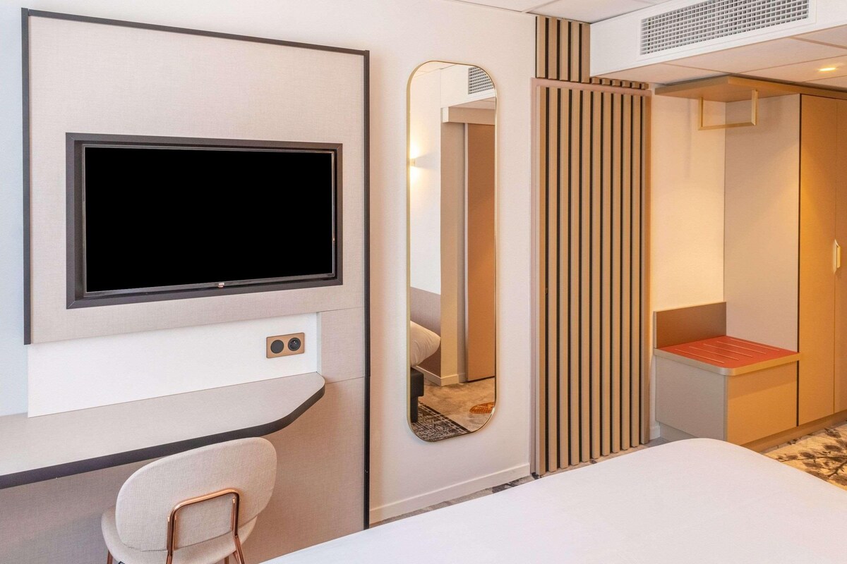 Ein Hotelzimmer mit einem an der Wand befestigten Flachbildfernseher, einem Spiegel, einem Bett, einem Sessel und einem Kleiderschrank.