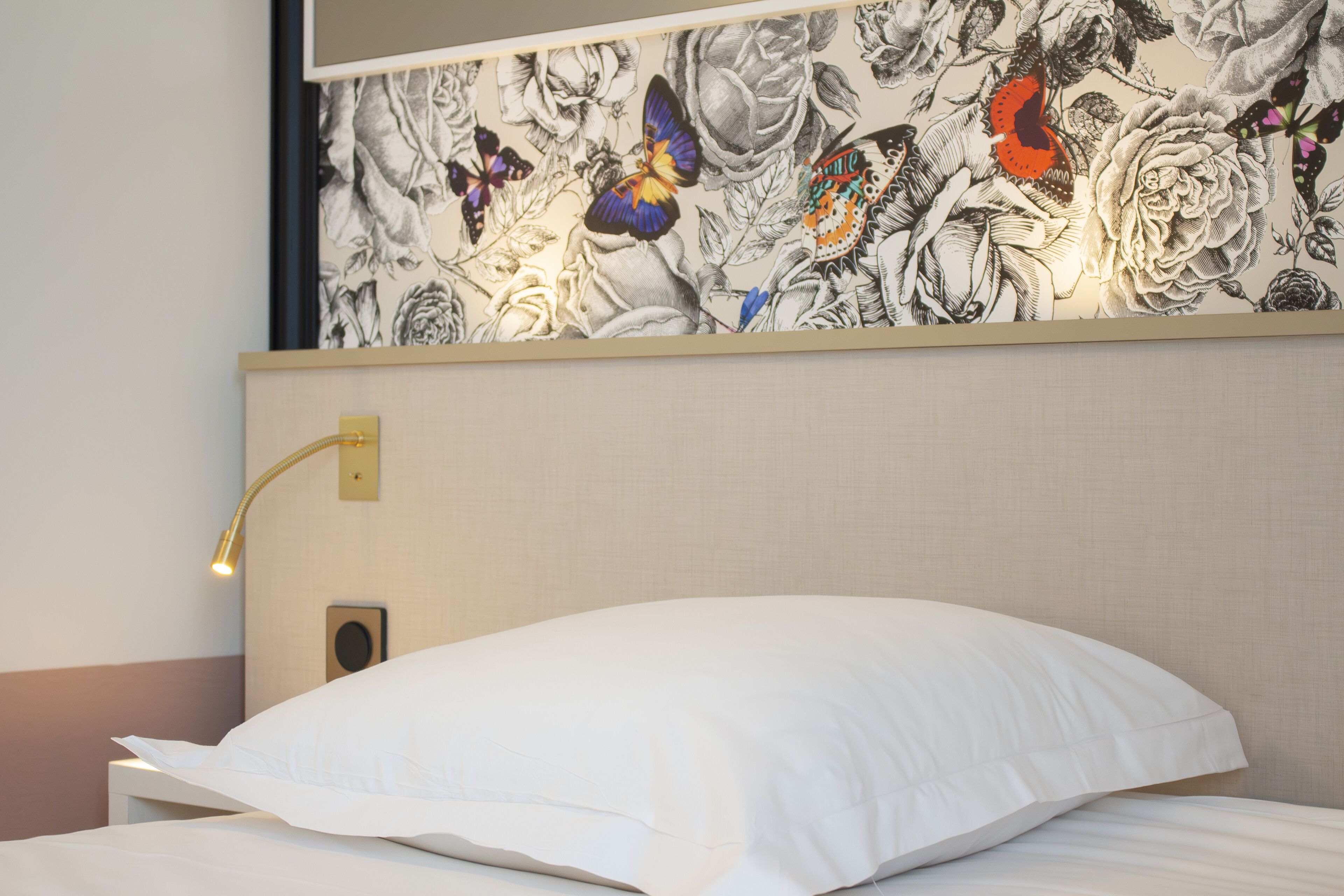 Une chambre d’hôtel avec un lit, une lampe fixée au mur et un papier peint à motifs floraux.