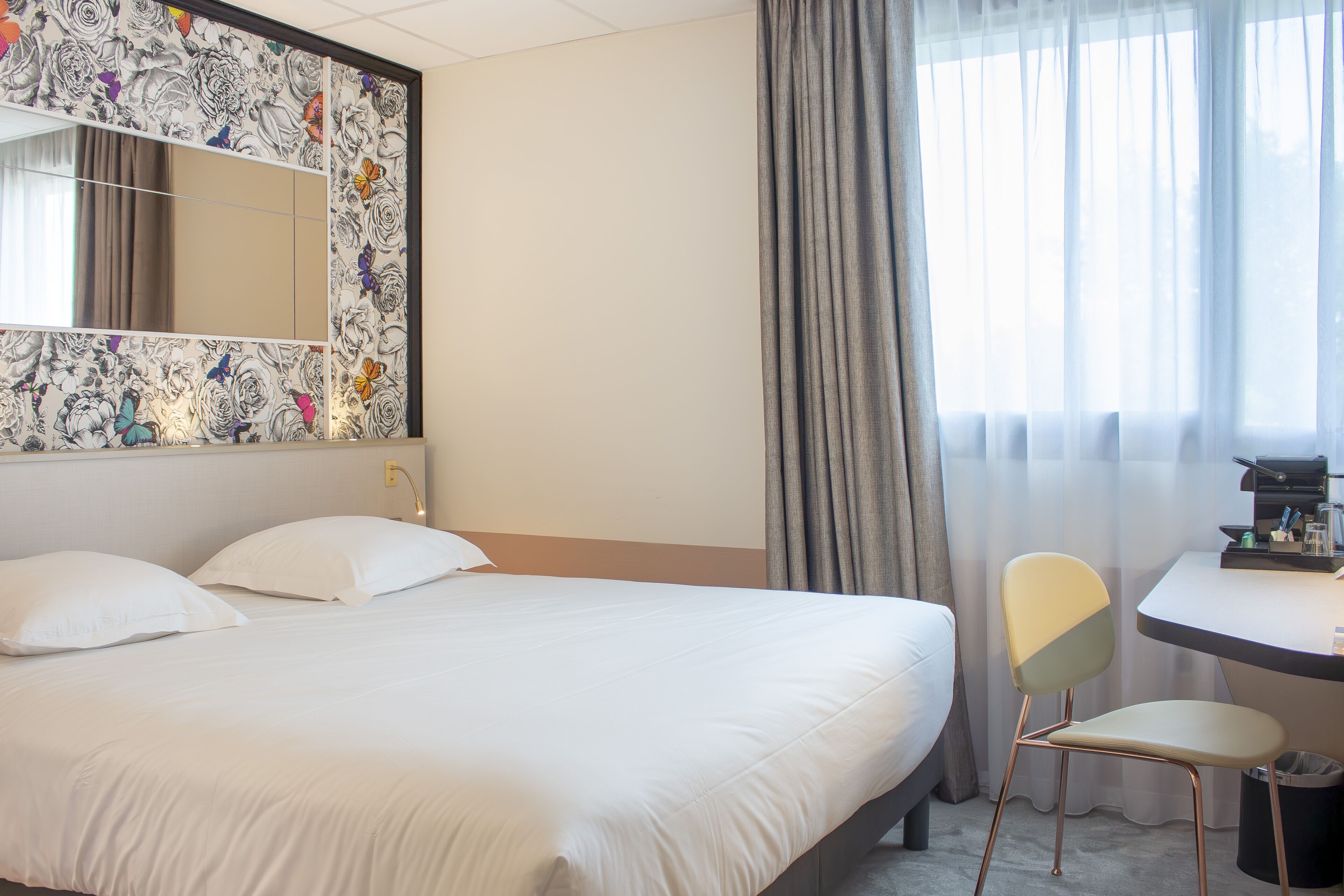 A hotel room with a bed, a chair, a desk, a mirror, and a window with curtains.
