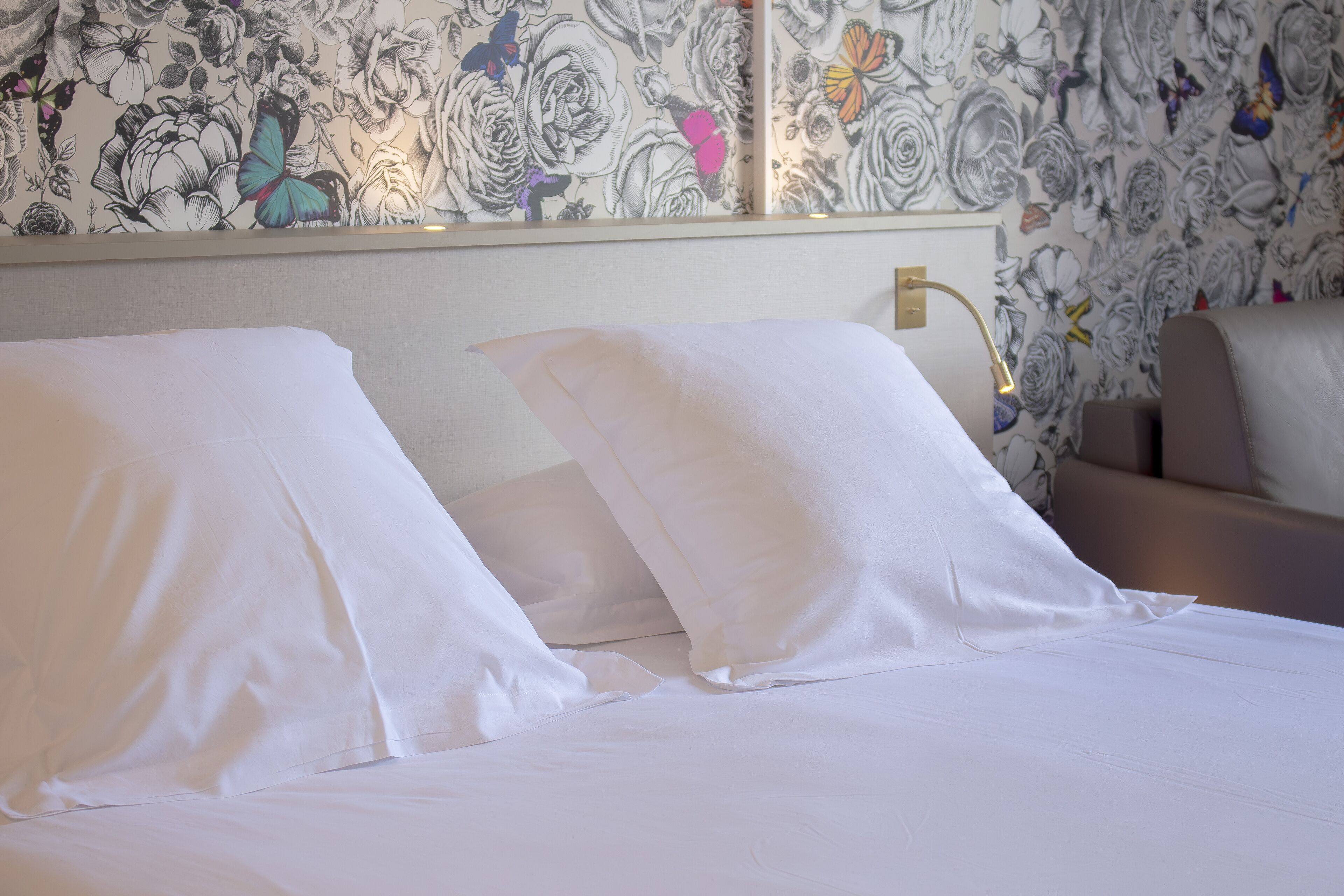 Una habitación de hotel con una cama, almohadillas blancas, una mesita de noche, una lámpara y un papel tapiz floral.