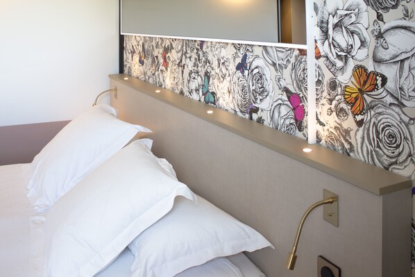Une chambre d’hôtel avec un lit, des oreillers blancs, une tête de lit à motifs floraux, un miroir et un luminaire fixé au mur.