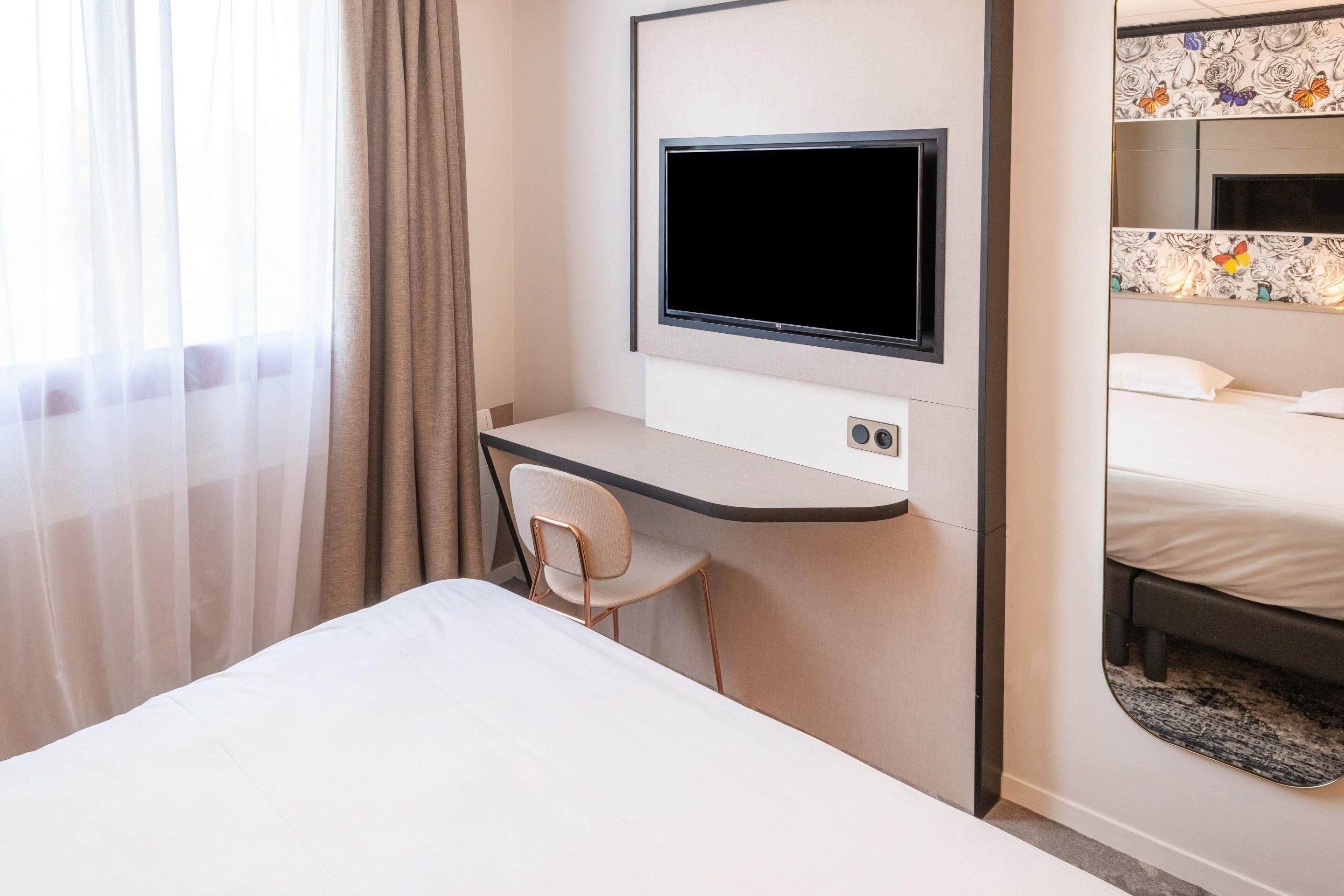 Une chambre d’hôtel comprenant un lit, un bureau avec une chaise, un téléviseur fixé au mur et un miroir au-dessus du lit.