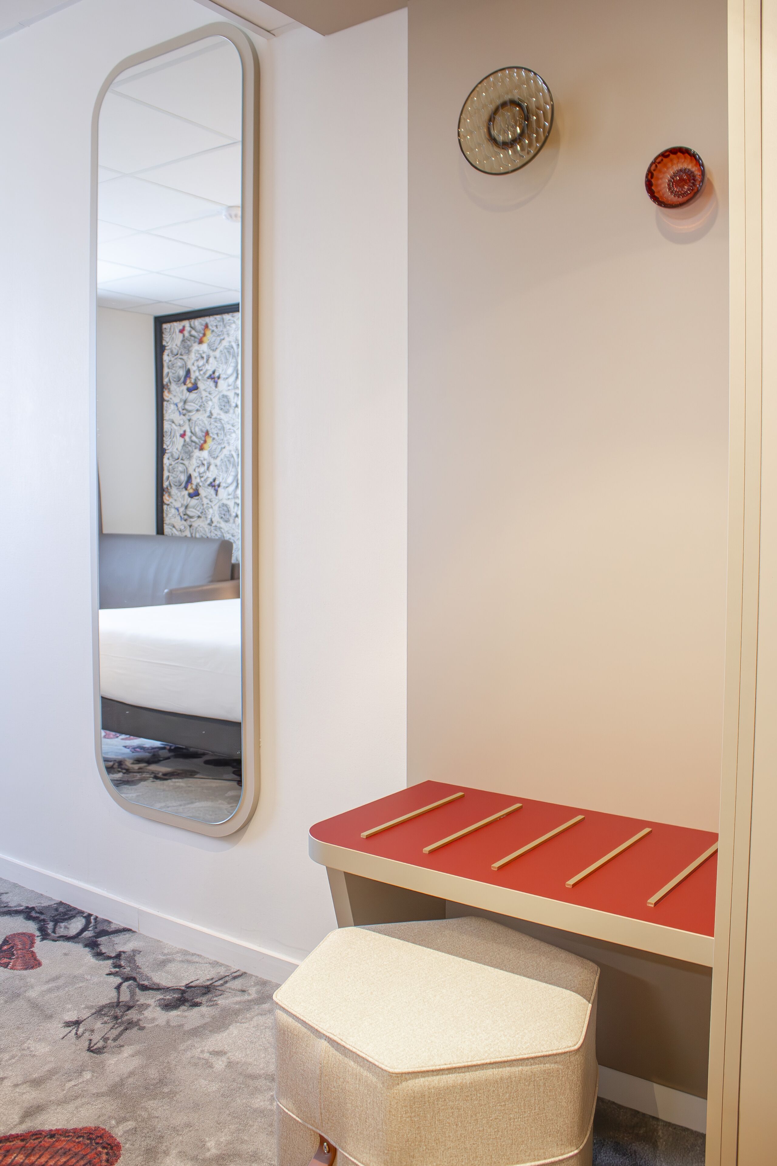 A hotel room with a large mirror, a bed, a red bench, and a small stool.