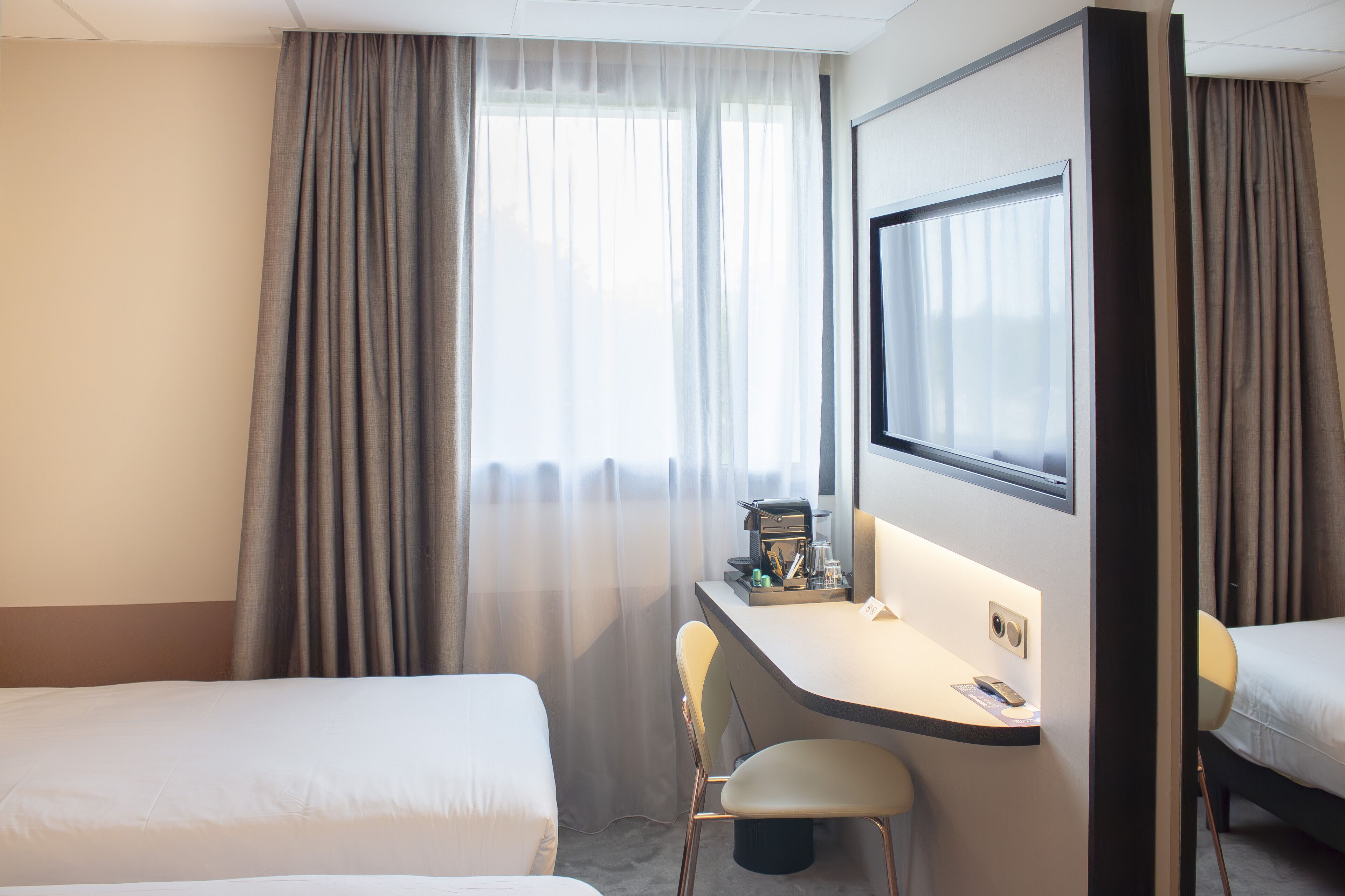 A hotel room with two beds, a desk with a chair, a large mirror, and a window with curtains.