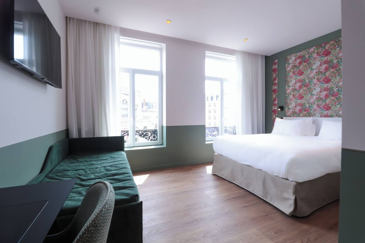 A hotel room with a bed, a desk, and a chair. There are two windows with curtains, and a balcony is visible through the window.