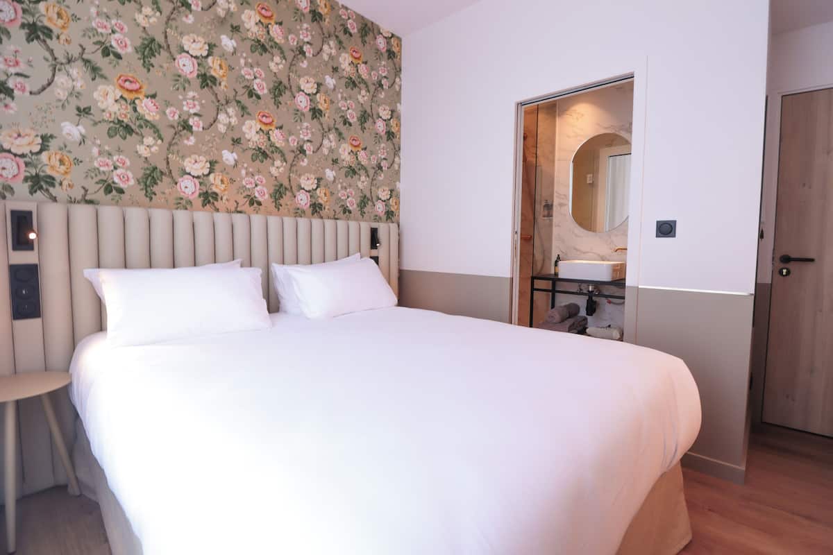 A bedroom with a large bed, a headboard, a floral wallpaper, a wooden door, and a bathroom visible through an open door.