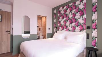 Une chambre à coucher avec un lit, un miroir, un mur recouvert d’un papier peint à motifs floraux et une porte.