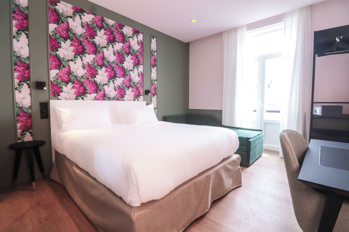 A hotel room with a large bed, a green accent wall, a small side table, a green ottoman, a desk with a chair, and a window with curtains.
