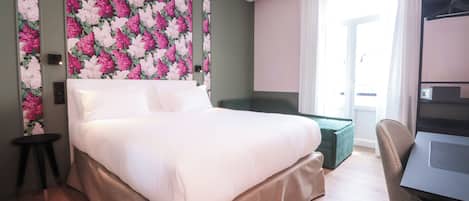 Habitación de hotel con una cama grande, una pared de acento verde, una mesita de noche pequeña, un otoman verde, un escritorio con silla y una ventana con cortinas.