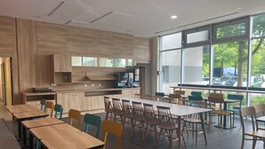 A modern dining area with wooden tables and chairs, a kitchen counter, and large windows offering a view of the outdoors.