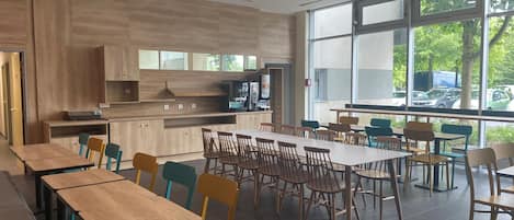 Uma área de refeições moderna com mesas e cadeiras de madeira, bancada de cozinha e janelas amplas com vista para o exterior.