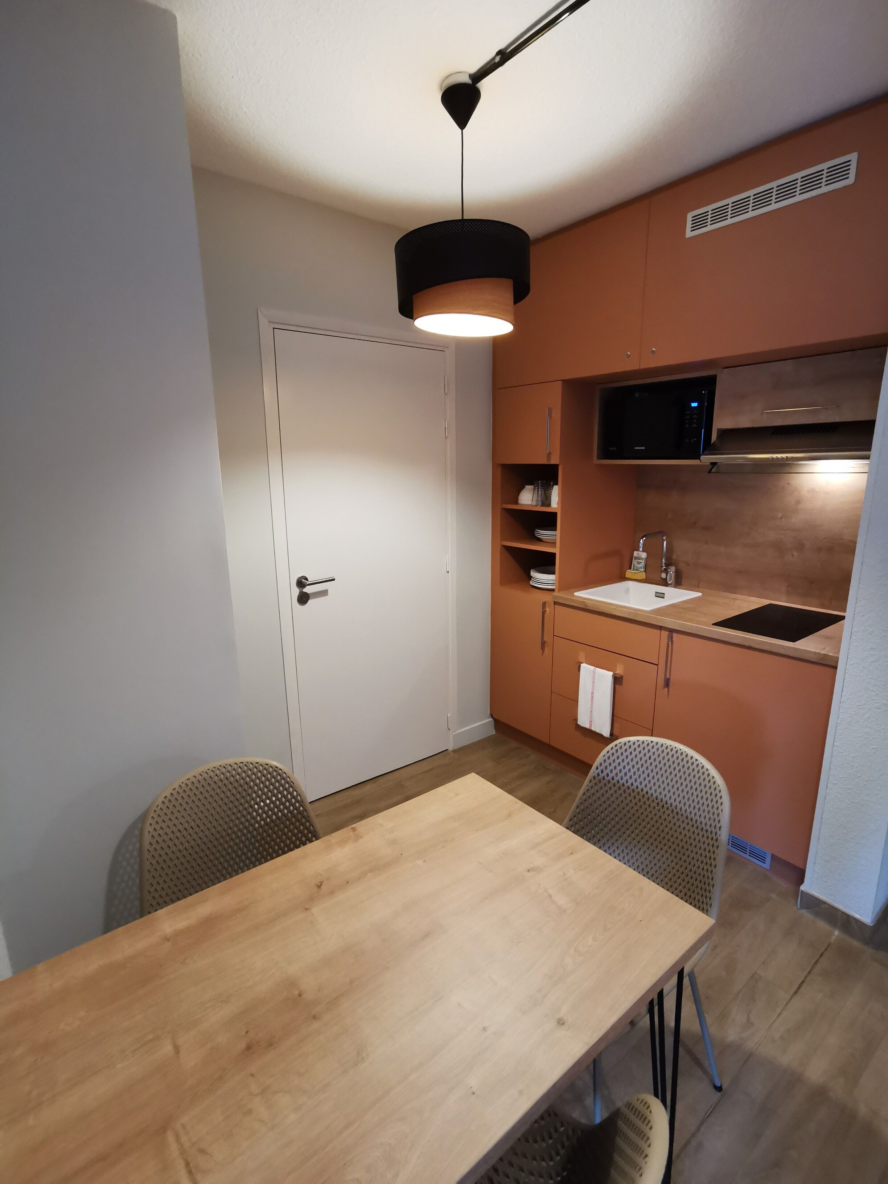 Una cocina compacta con microondas, lavamanos y armarios de madera. Hay una mesa de comedor con dos sillas.