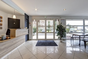 Uma área de recepção moderna com balcão de madeira, televisão, planta em vaso e vista para o estacionamento através de portas de vidro.