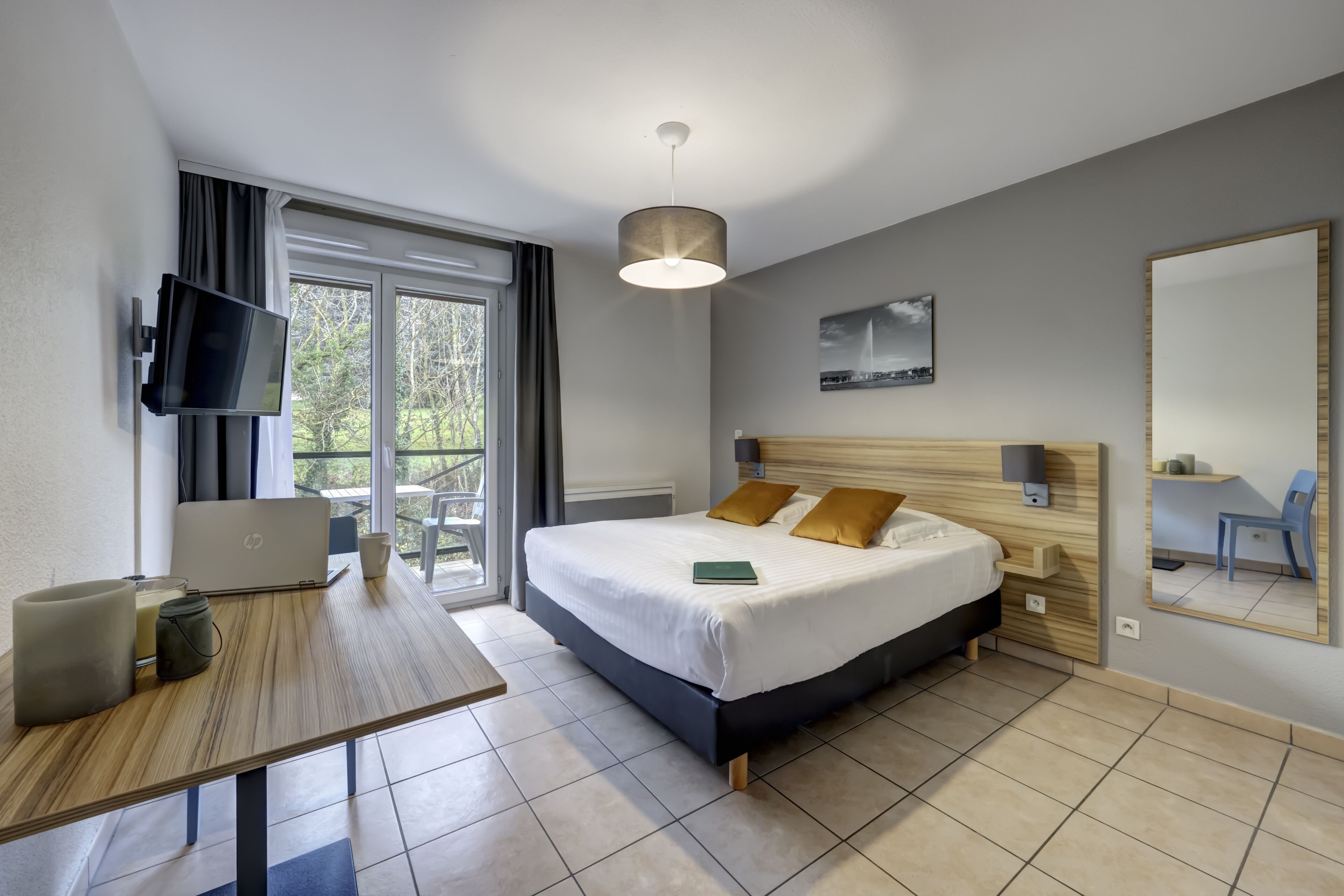 Una habitación de hotel moderna con una cama, un escritorio con una computadora portátil, un televisor y un ventanal grande.