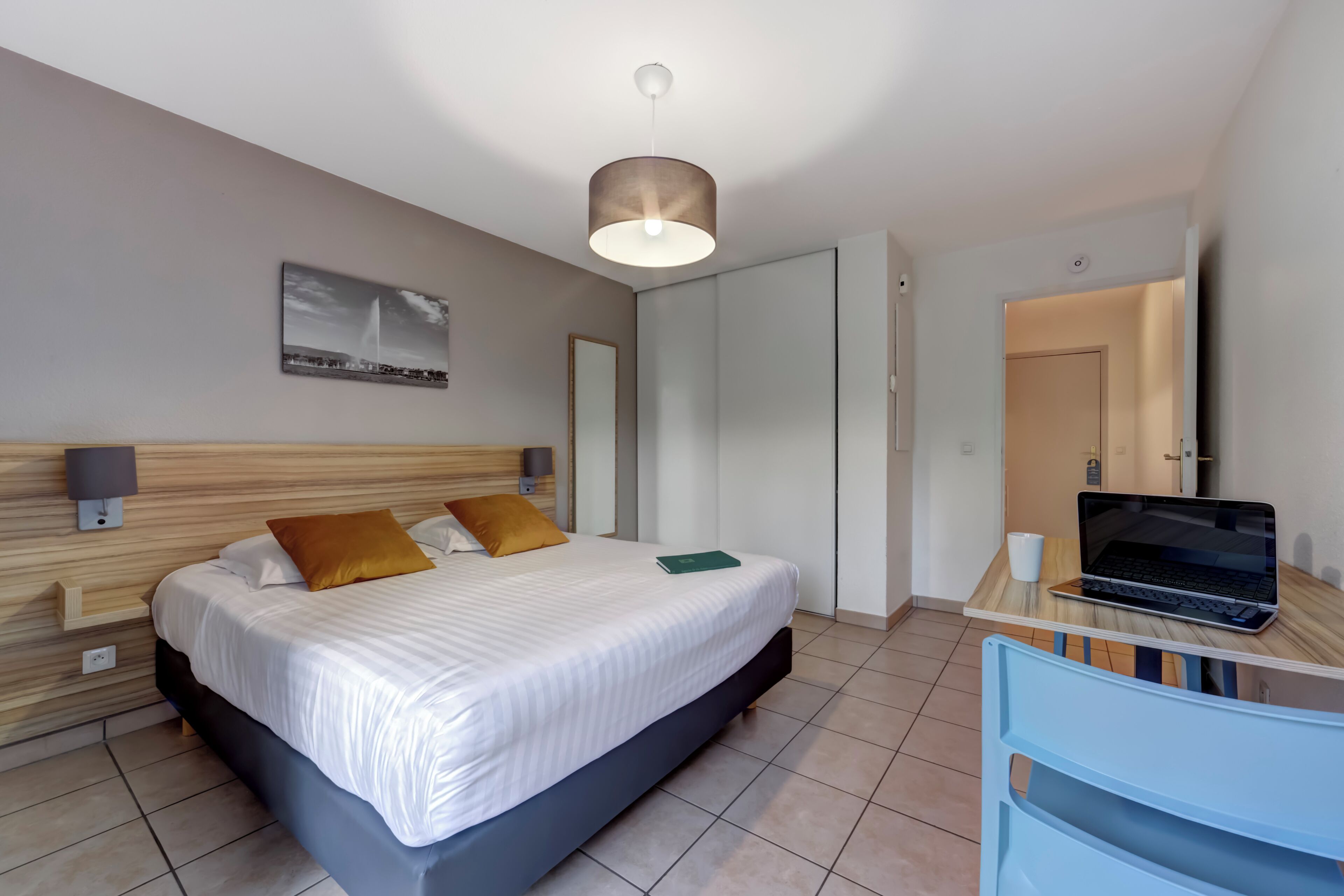 A modern hotel room with a bed, bedside lamps, a desk with a laptop, and a painting on the wall.