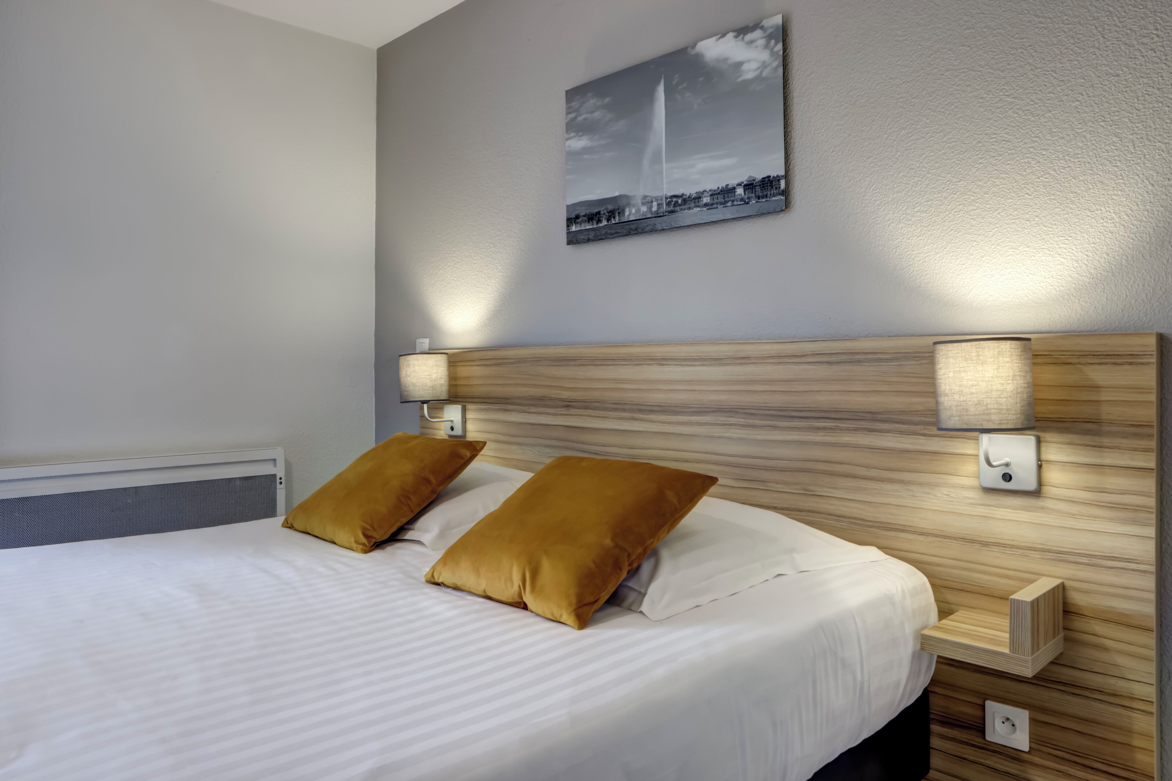 Een net opgemaakt bed met twee kussens, een hoofdbord met ingebouwde verlichting, een klein nachtkastje en een aan de muur hangend schilderij.