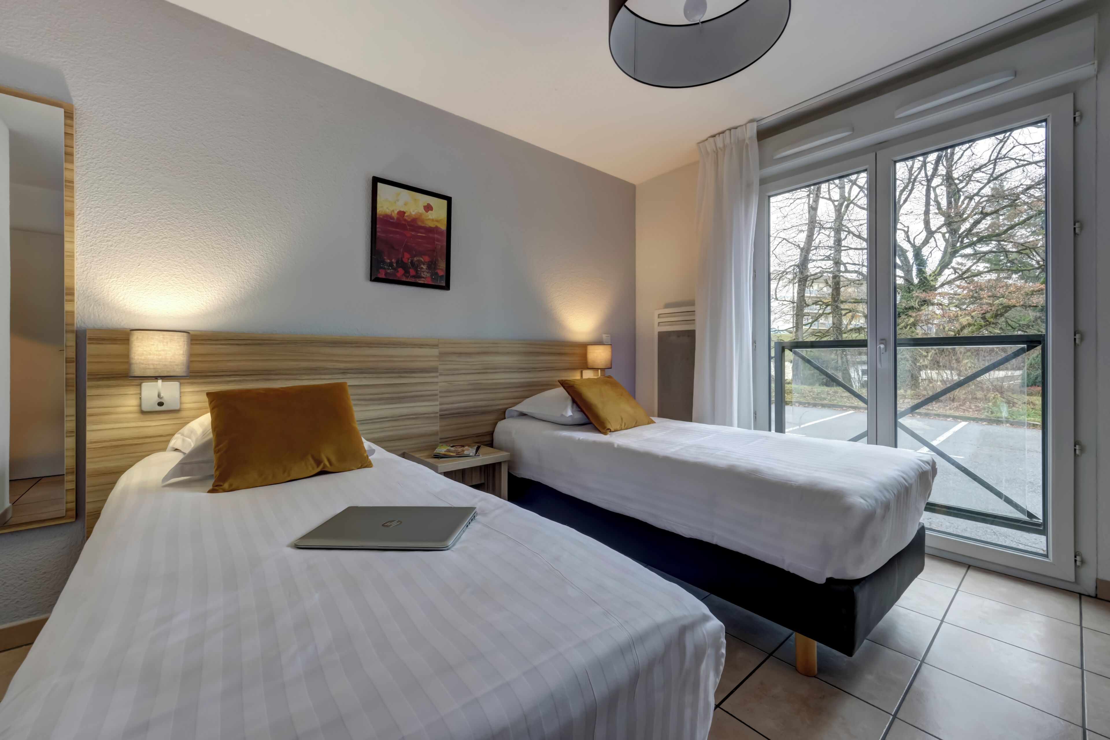 A hotel room with two beds, a painting on the wall, a laptop on one bed, and a view of the outside through the window.