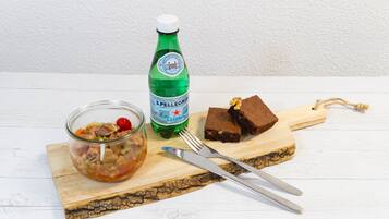A wooden cutting board with a glass jar of salad, a bottle of Pellegrino water, and a piece of chocolate cake.