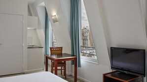 Hotelkamer met een bed, een televisie, een eettafel met stoel, een kitchenette en uitzicht op gebouwen door een raam.