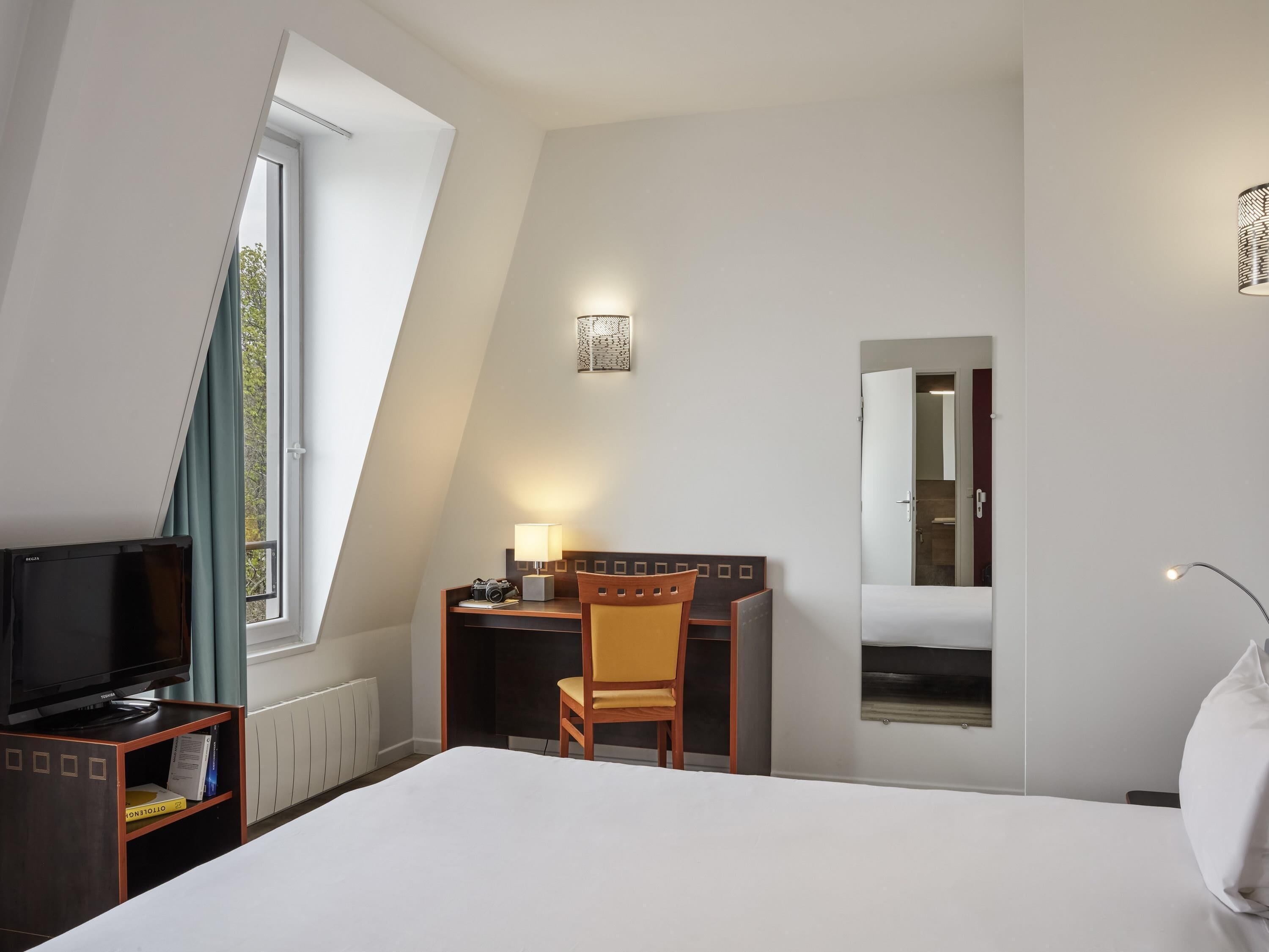 Habitación de hotel con cama, escritorio, silla, televisión, lámpara y ventana con cortinas.