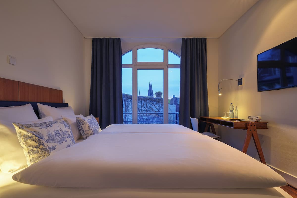 A hotel room with a large bed, a desk with a chair, a TV on the wall, and a window with curtains.