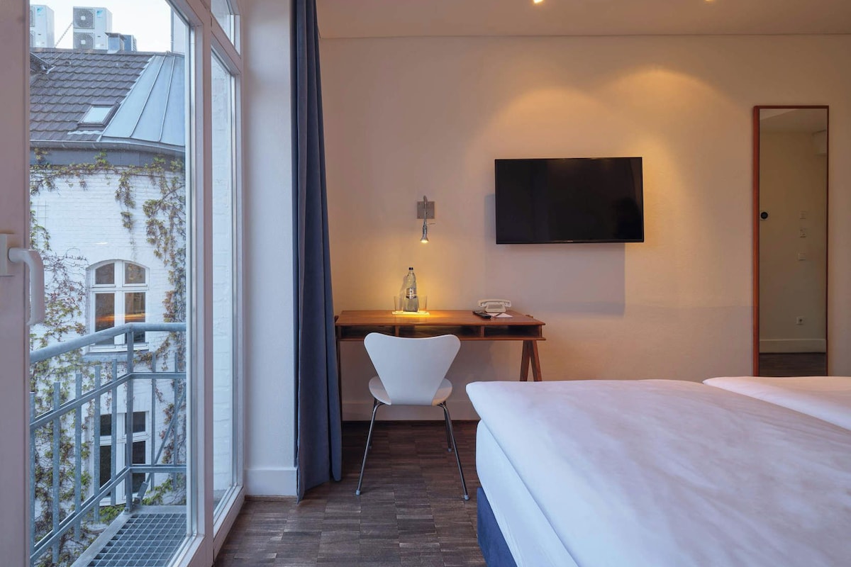 Une chambre d’hôtel comprenant un lit, un bureau avec une chaise, une télévision et une fenêtre donnant sur des immeubles.