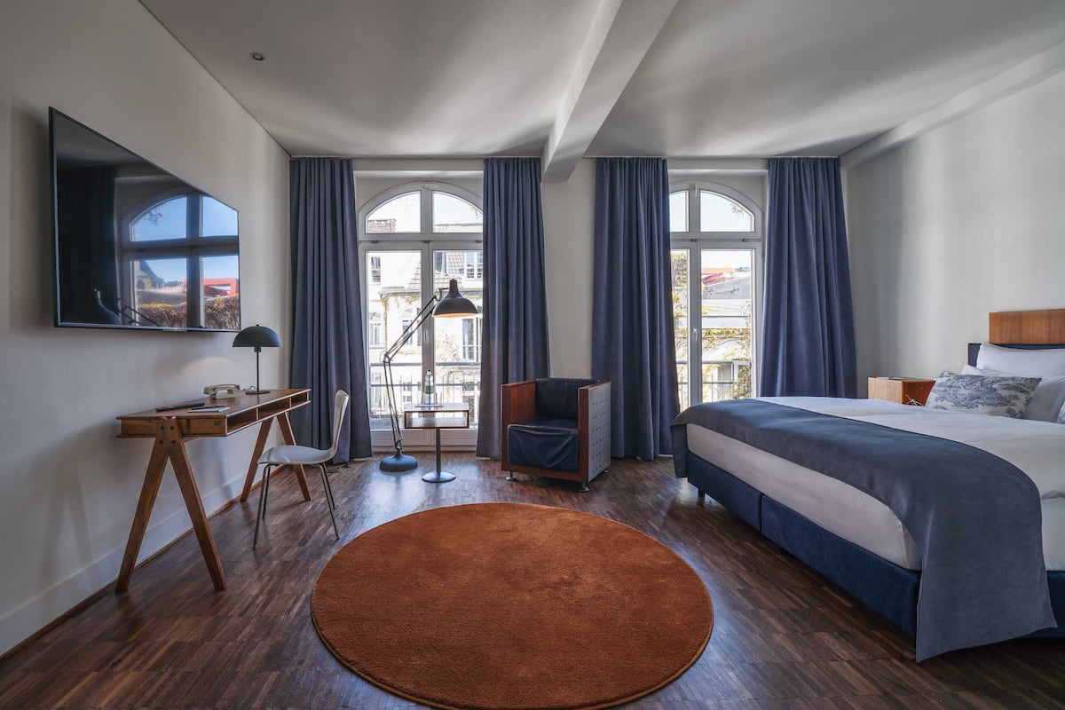 A modern hotel room with a large bed, a desk with a chair, a round rug, and a large window with curtains.