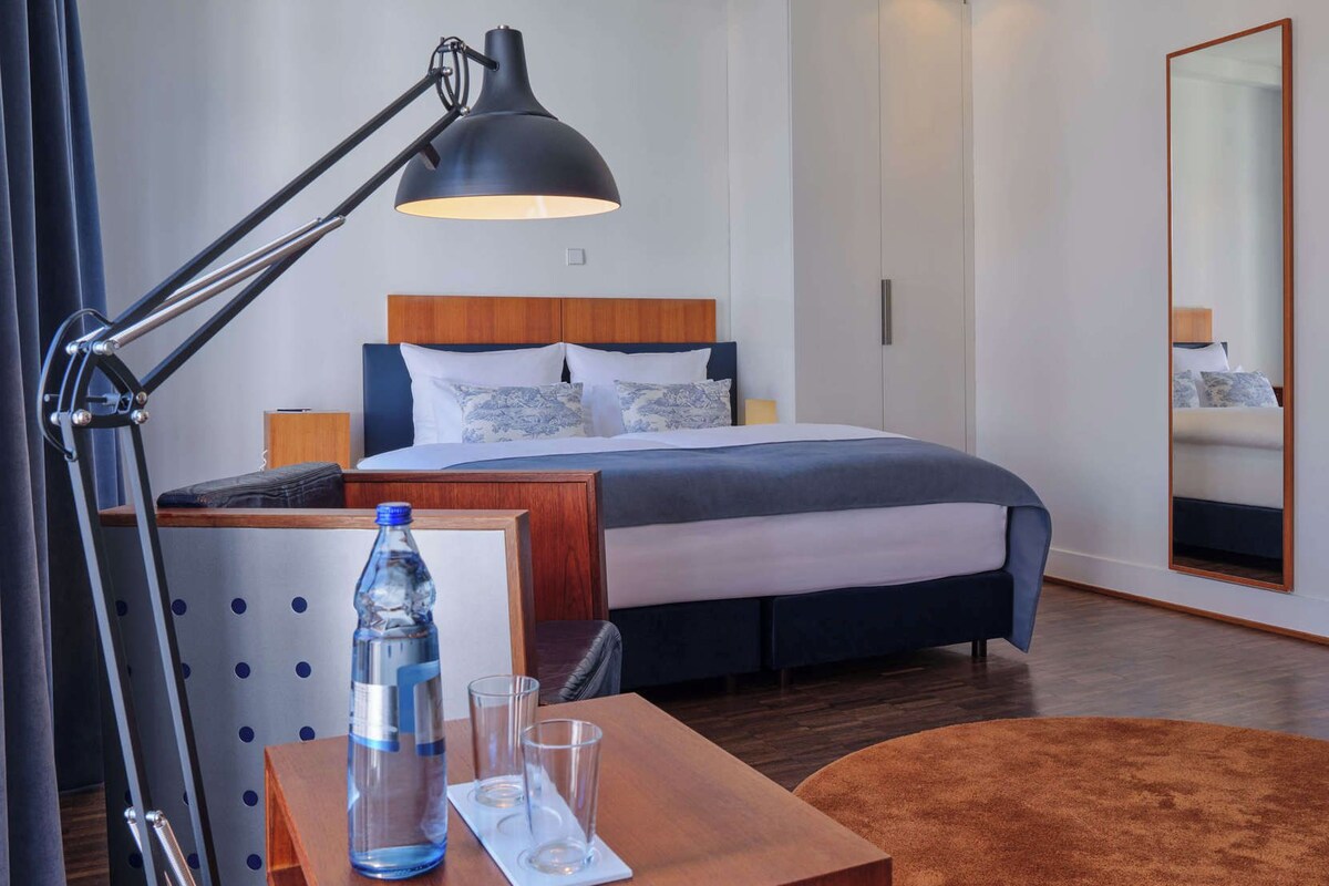 Une chambre d’hôtel avec un lit, un bureau doté d’une lampe, une bouteille d’eau et des verres.