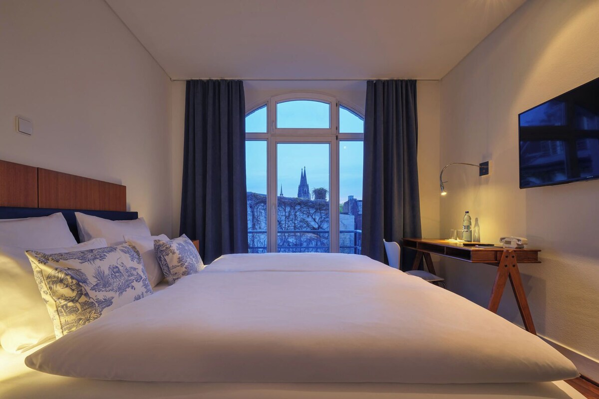 A hotel room with a large bed, a desk with a chair, a TV, and a view of a cityscape through a window.