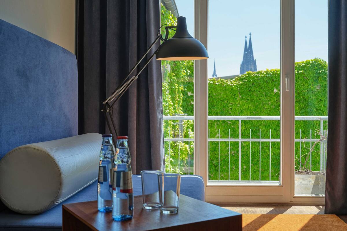 A hotel room with a sofa, a table with water bottles and glasses, a lamp, and a view of a church spire through a balcony.