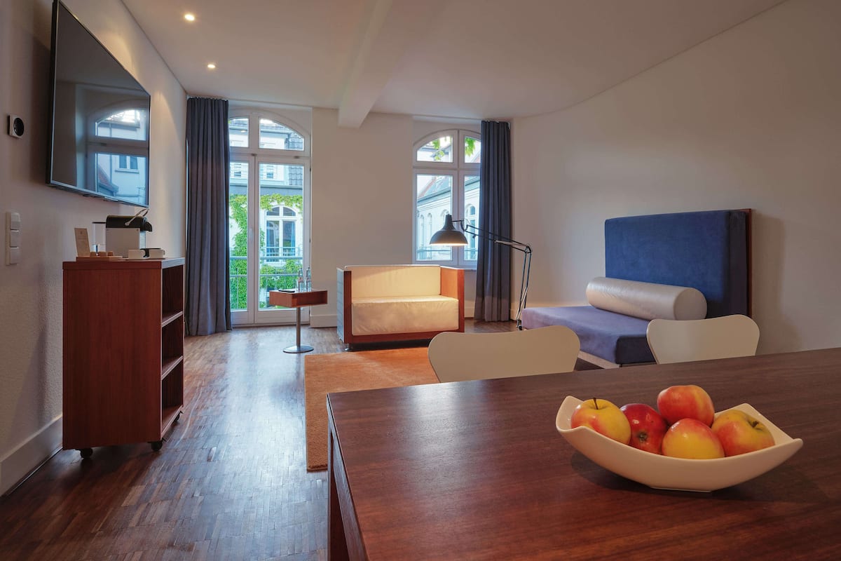 A modern hotel room with a wooden floor, a dining table with a bowl of fruit, a sofa, a bed, a TV, and a window with a view of buildings.