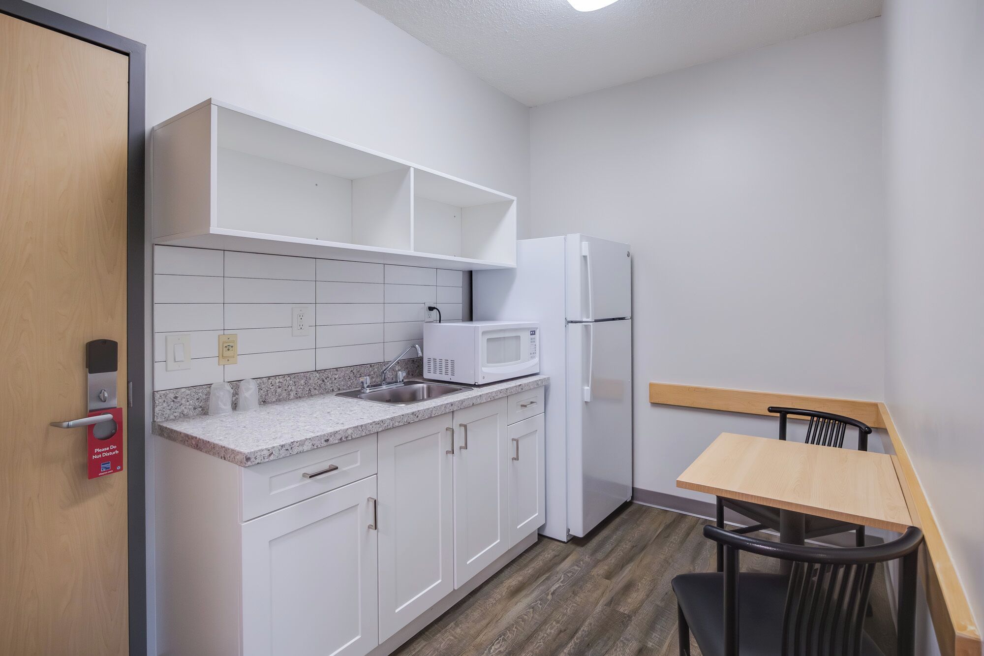Cocina compacta con armarios blancos, encimera de granito, microondas y refrigerador. Hay una pequeña mesa y una silla en la esquina.