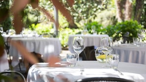 Uma área de refeições ao ar livre com mesas redondas cobertas por toalhas brancas, cada uma com copos de vinho, copos de água e talheres.