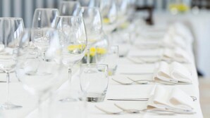 Uma mesa comprida posta com vários copos de vinho e pratos, provavelmente para um evento gastronômico formal.