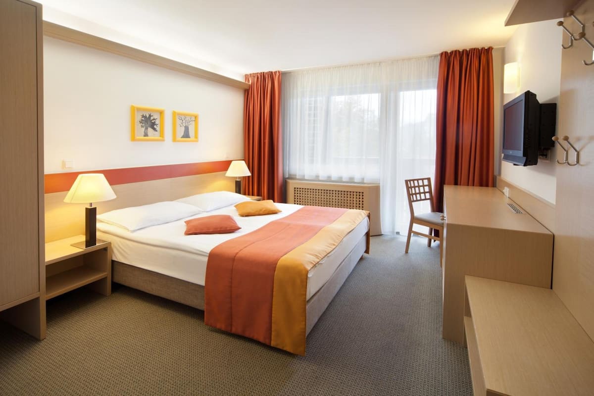 A hotel room with a bed, bedside table, lamp, TV, and a desk with a chair.
