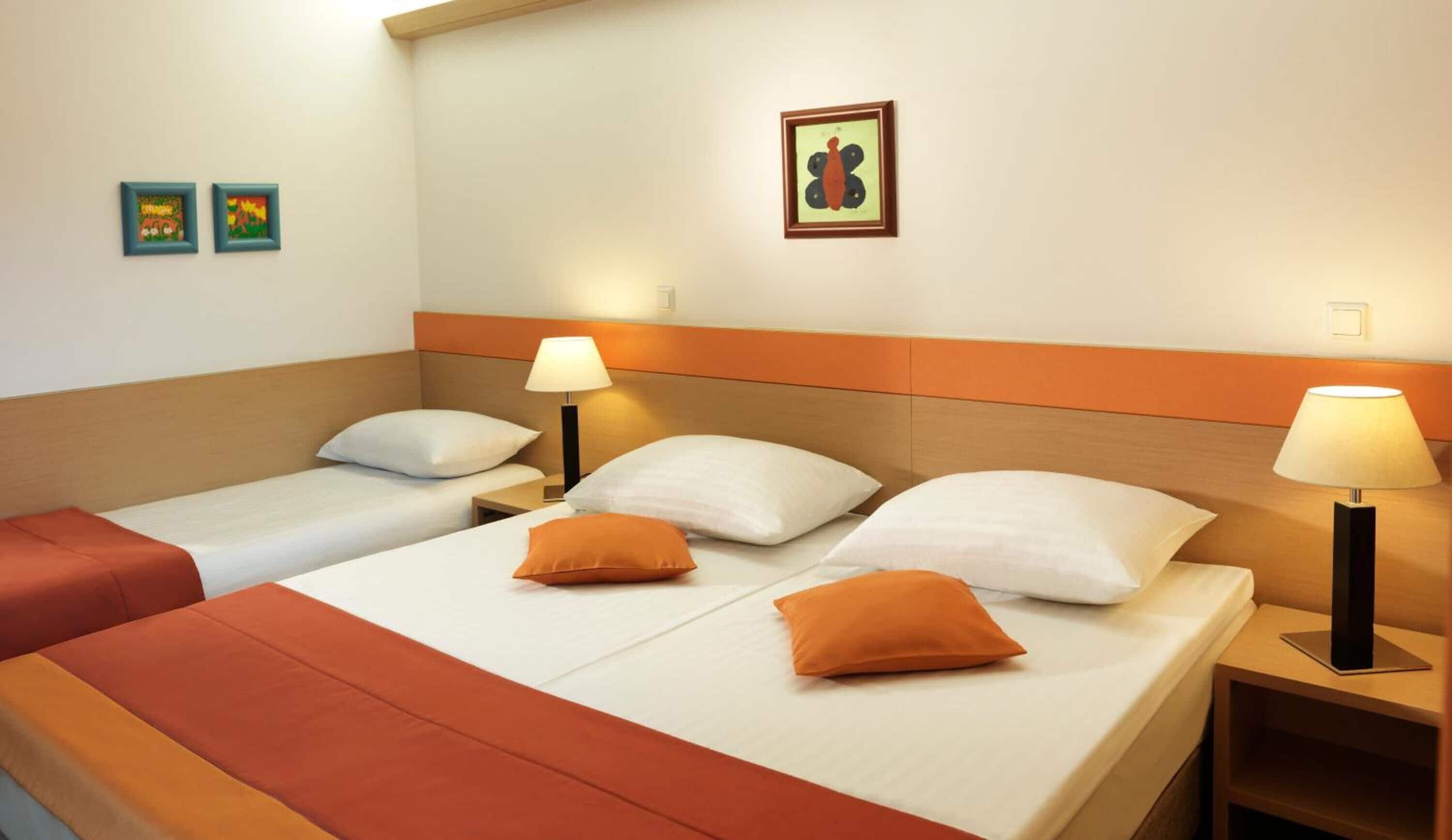 Camera d'albergo con due letti, ognuno con biancheria bianca e cuscini arancioni. Sono presenti due comodini con lampade e un quadro appeso al muro.